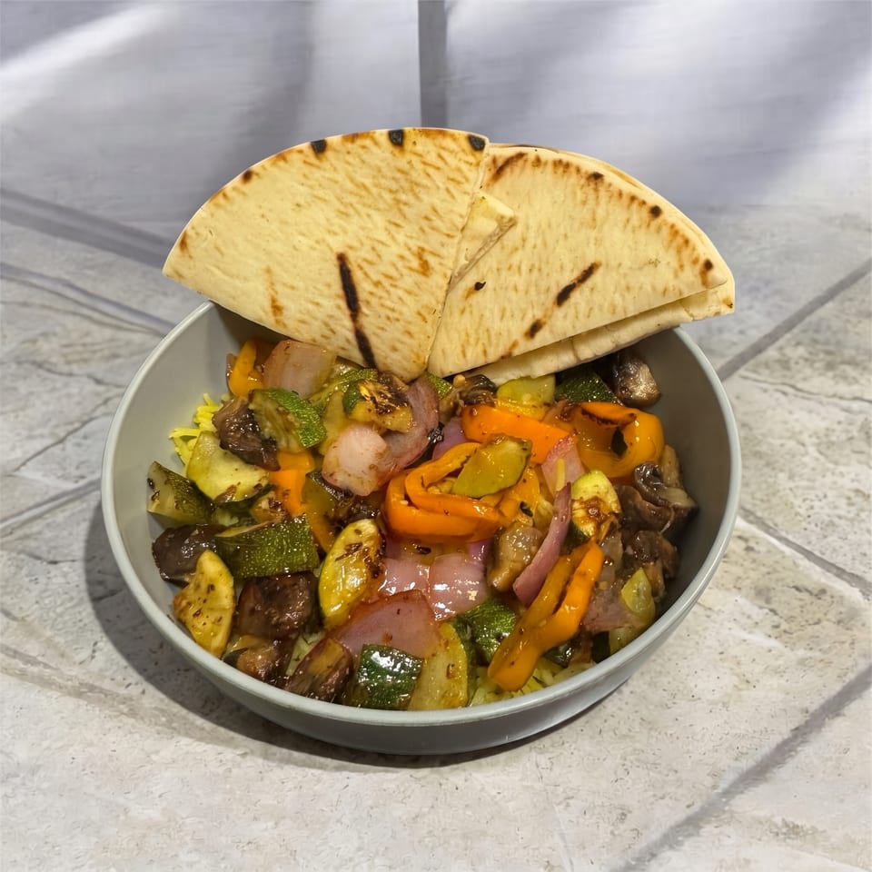 A bowl of grilled vegetables including zucchini, bell peppers, and red onion, topped with pita bread.